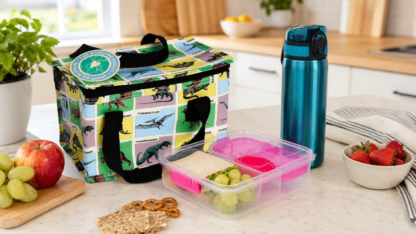 Dinosaur-print insulated lunch bag beside a teal water bottle and a pink compartment lunch box containing a sandwich and fruit on a bright kitchen worktop.